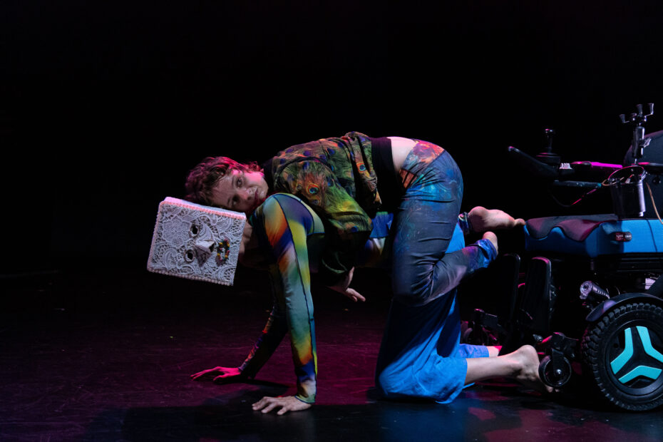 A performer wearing a robot head crawls along, while another performer clings to their back. Both performers look at the camera. A wheelchair is visible in the background.